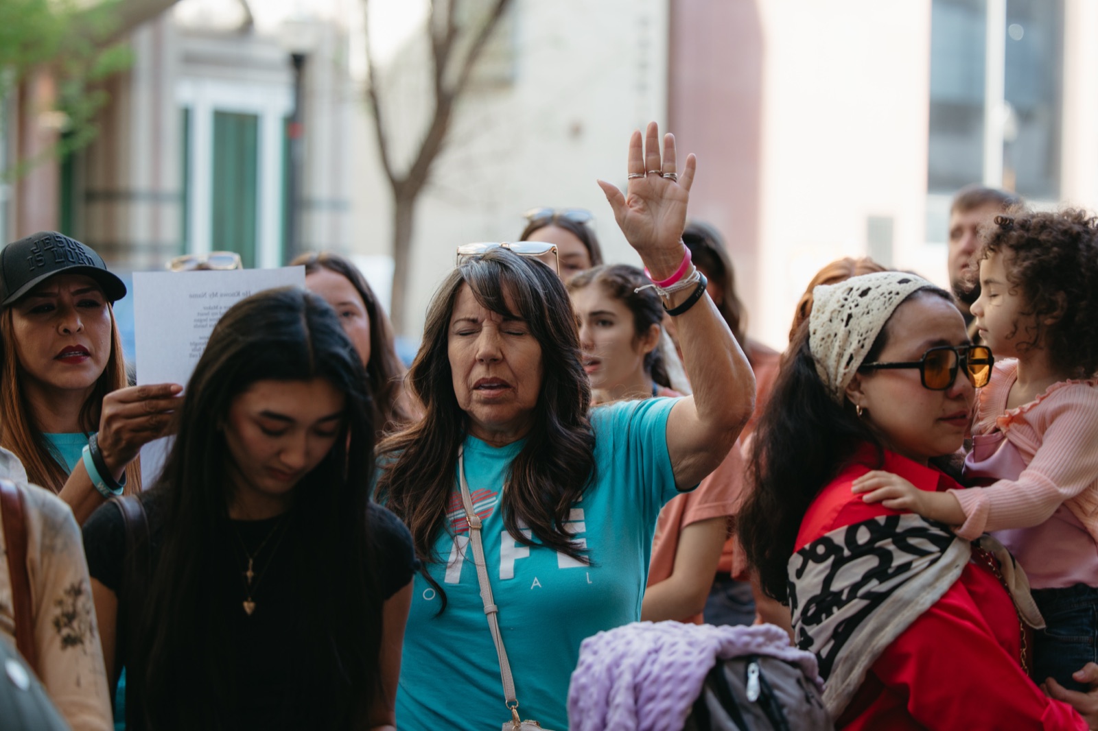 Women pray outdoors at a gathering, one raising her hand with eyes closed, another holding a child.